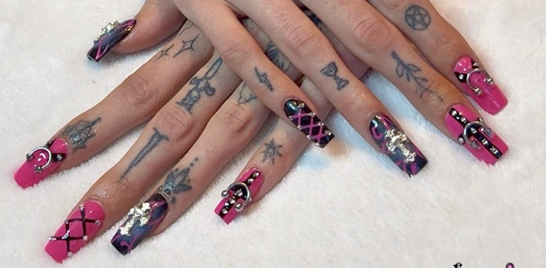 Close-up of hands with pink and black nail art on a white surface, featuring tattoos.