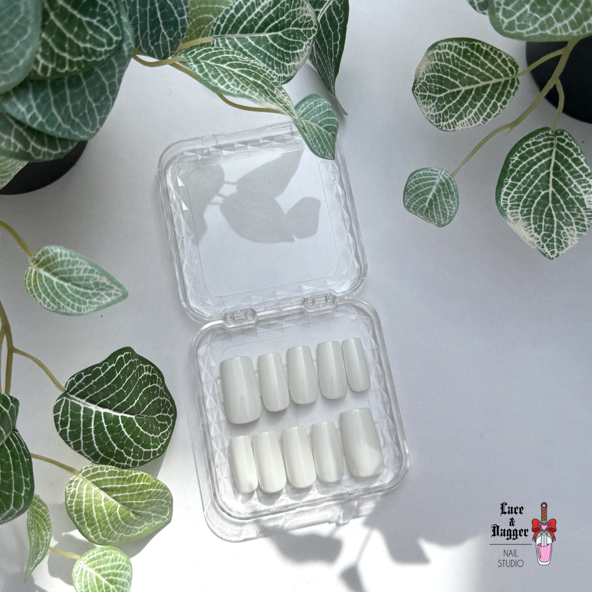 Set of Square handmade custom white press-on nails in a clear plastic case with green leafy plants in the background