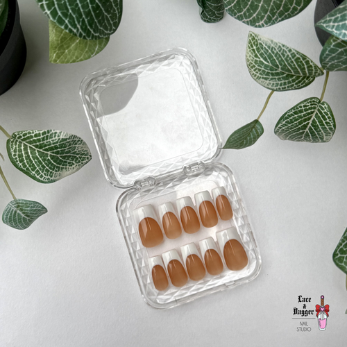 Set of square handmade custom dark nude deep french tip press-on nails in a clear plastic case on a white surface with green leaves.