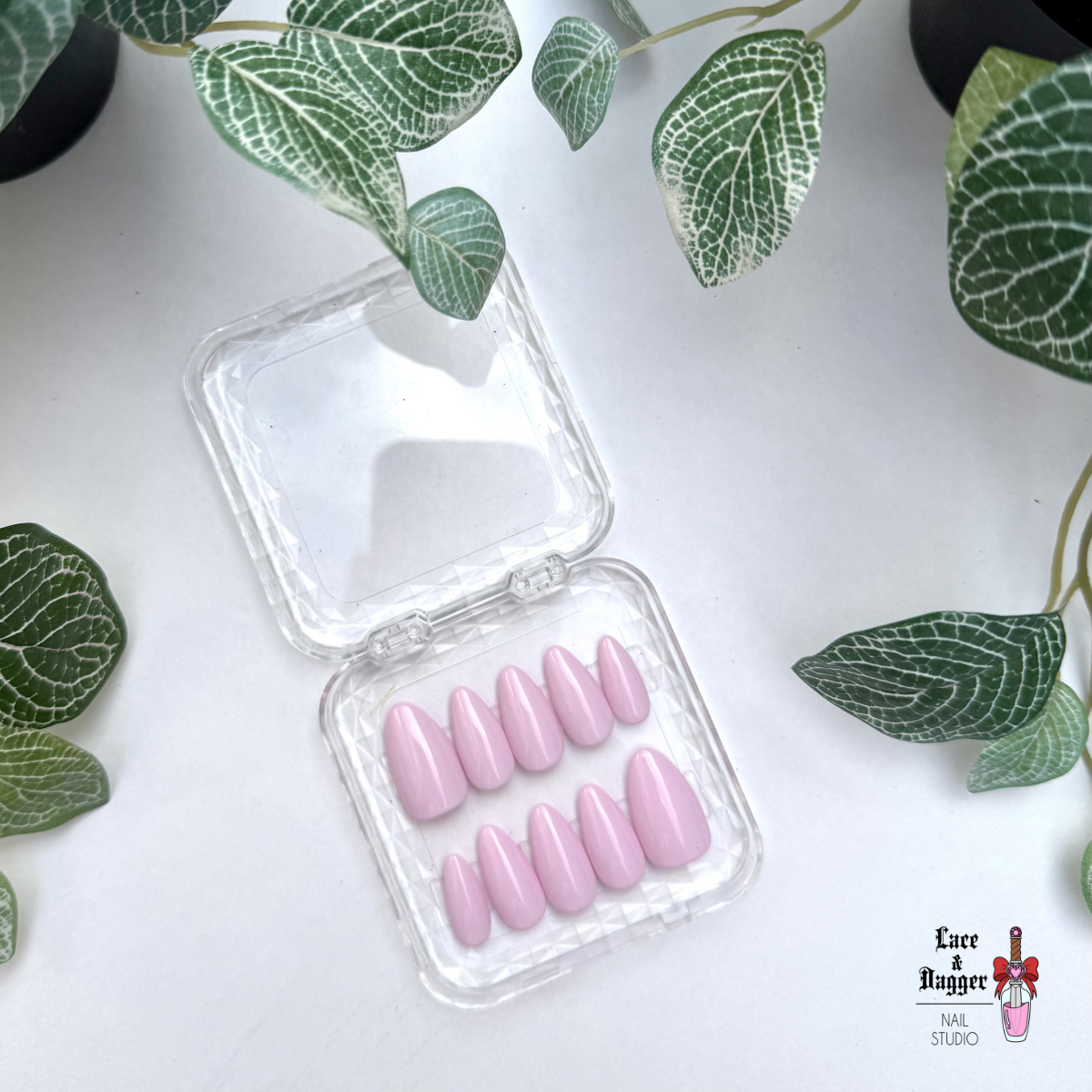 almond handmade custom pink press-on nails in a clear plastic case on a white background with green leaves.