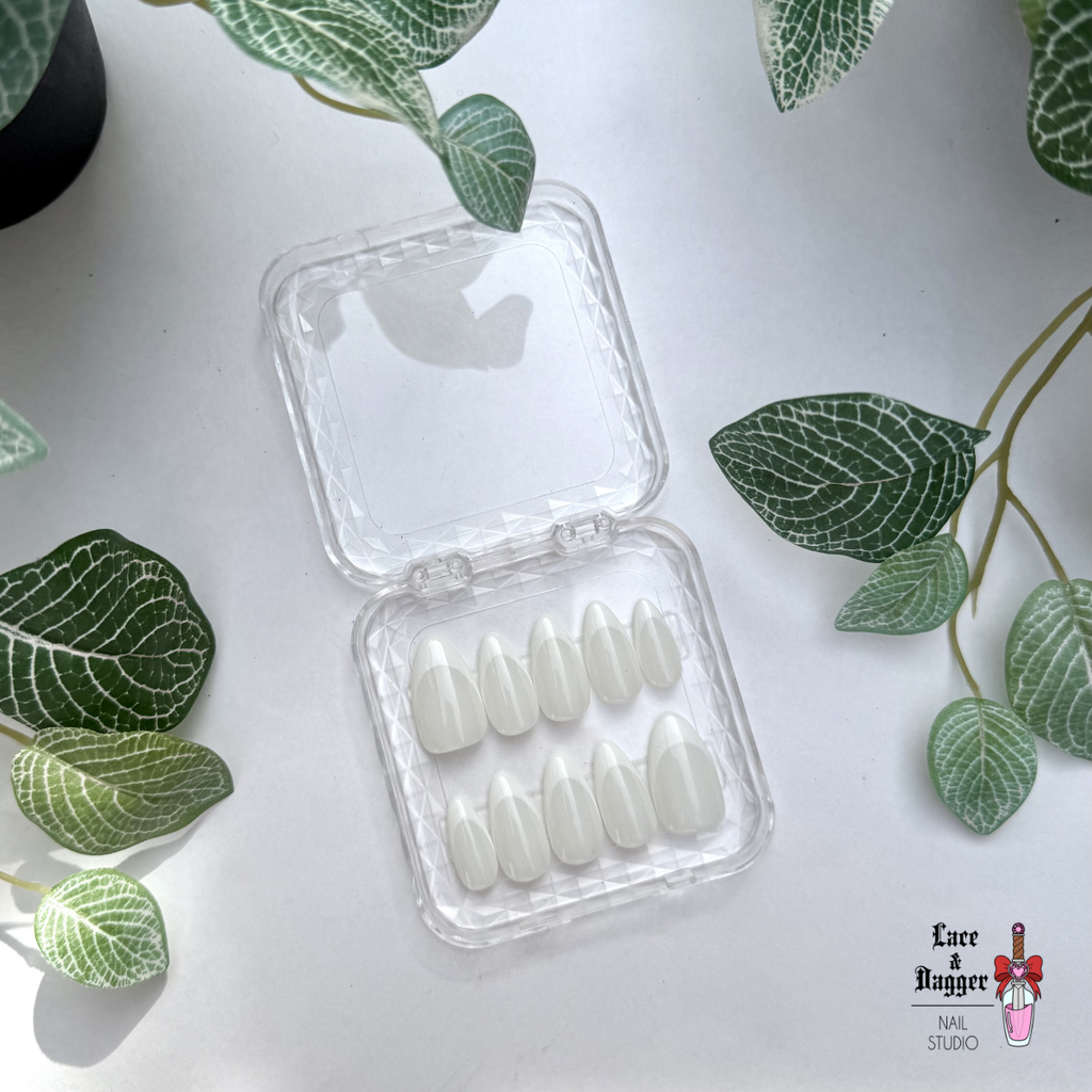 Set of handmade, custom almond milky-white deep French-tip press-on nails in a clear plastic case on a light surface with green leaves.