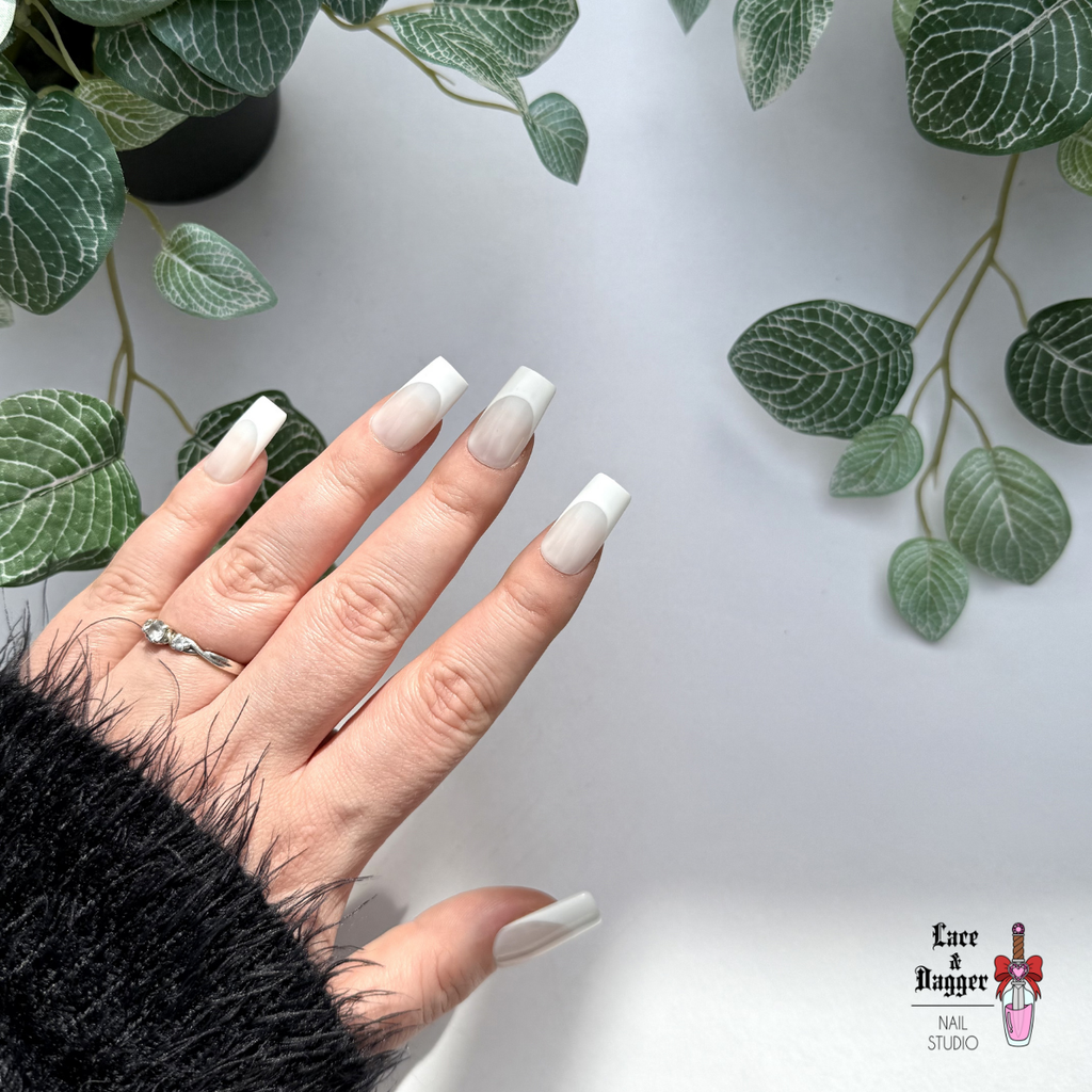 Hand with handmade custom square milky white deep french tip press on nails against a light background with green leaves