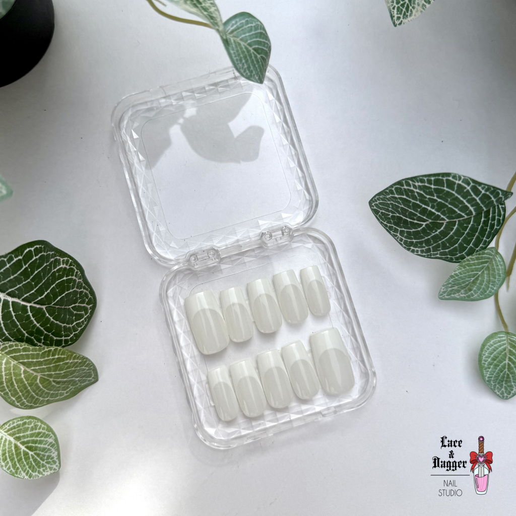 Set of handmade, custom square milky-white deep French-tip press-on nails in a clear plastic case on a light surface with green leaves.