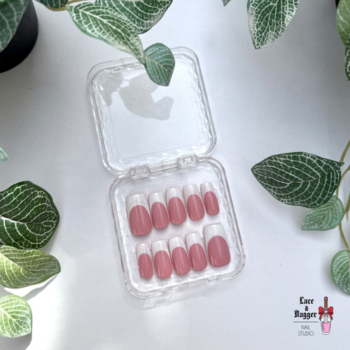 Set of square handmade custom dark pink deep french press on nails in a clear case on a light surface with green leaves.