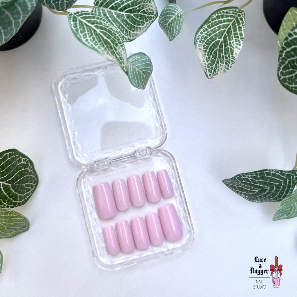 square handmade custom pink press on nails in an open clear container on a white background with green leaf decorations.