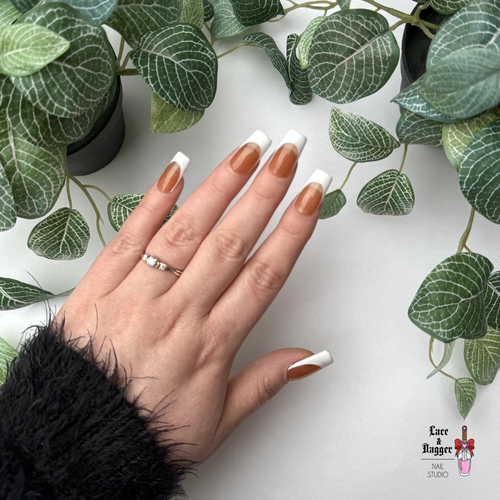 Hand with square homemade custom dark nude deep french tip press-on nails against a white background with green leaves.